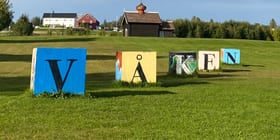 Store, fargerike bokstaver på treklosser som sammen utgjør "Våken". Står på en gressplen foran en gård.