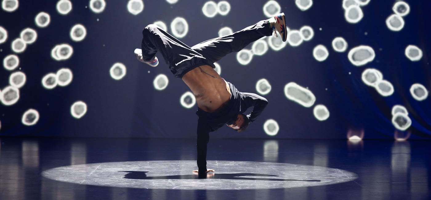 En hiphop-danser holder seg på gulvet med en hånd mens han danser opp-ned. Bak scenen ser det ut som om det er hvite rundinger.