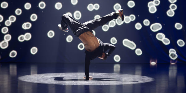 En hiphop-danser holder seg på gulvet med en hånd mens han danser opp-ned. Bak scenen ser det ut som om det er hvite rundinger.