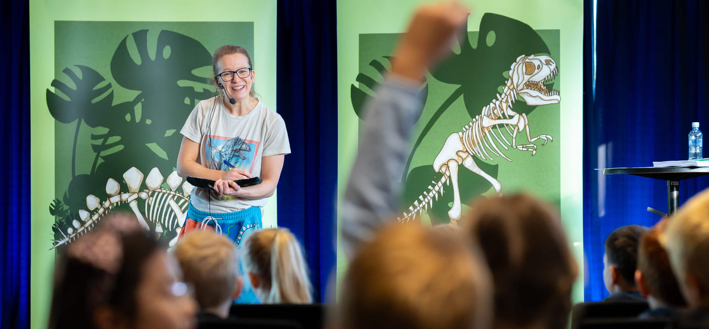 En kvinne står på en scene og snakker til en gruppe barn. Ett av barna rekker opp hånden.