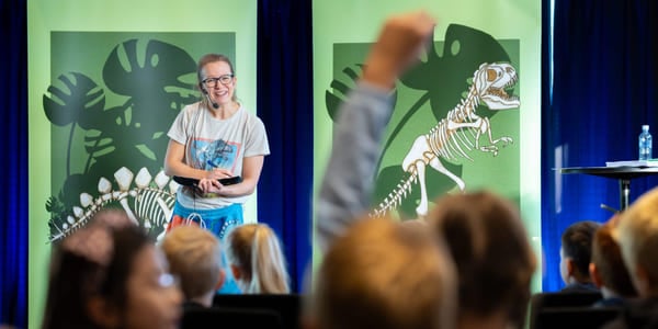 En kvinne står på en scene og snakker til en gruppe barn. Ett av barna rekker opp hånden.