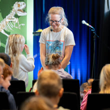 En kvinne står på scenen foran en gruppe barn, og smiler til en jente som står foran henne.