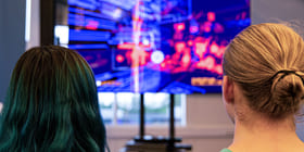 To personer sitter med ryggen til og ser på en skjerm med masse farger, antakelig et dataspill.