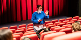 Skoleelever sitter i en kinosal og hører på en kvinne som står og snakker mellom to seterader.