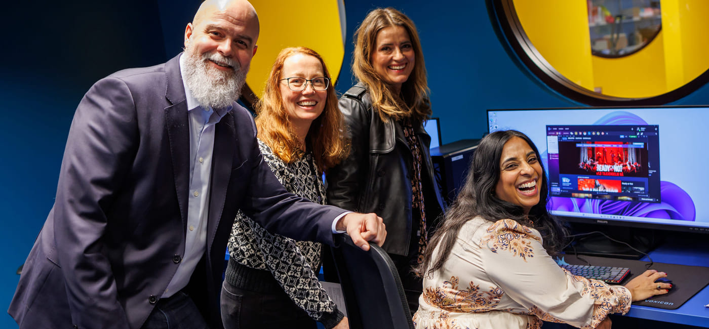 Fire personer smiler til kamera foran en pc-skjerm.