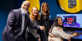 Fire personer smiler til kamera foran en pc-skjerm.