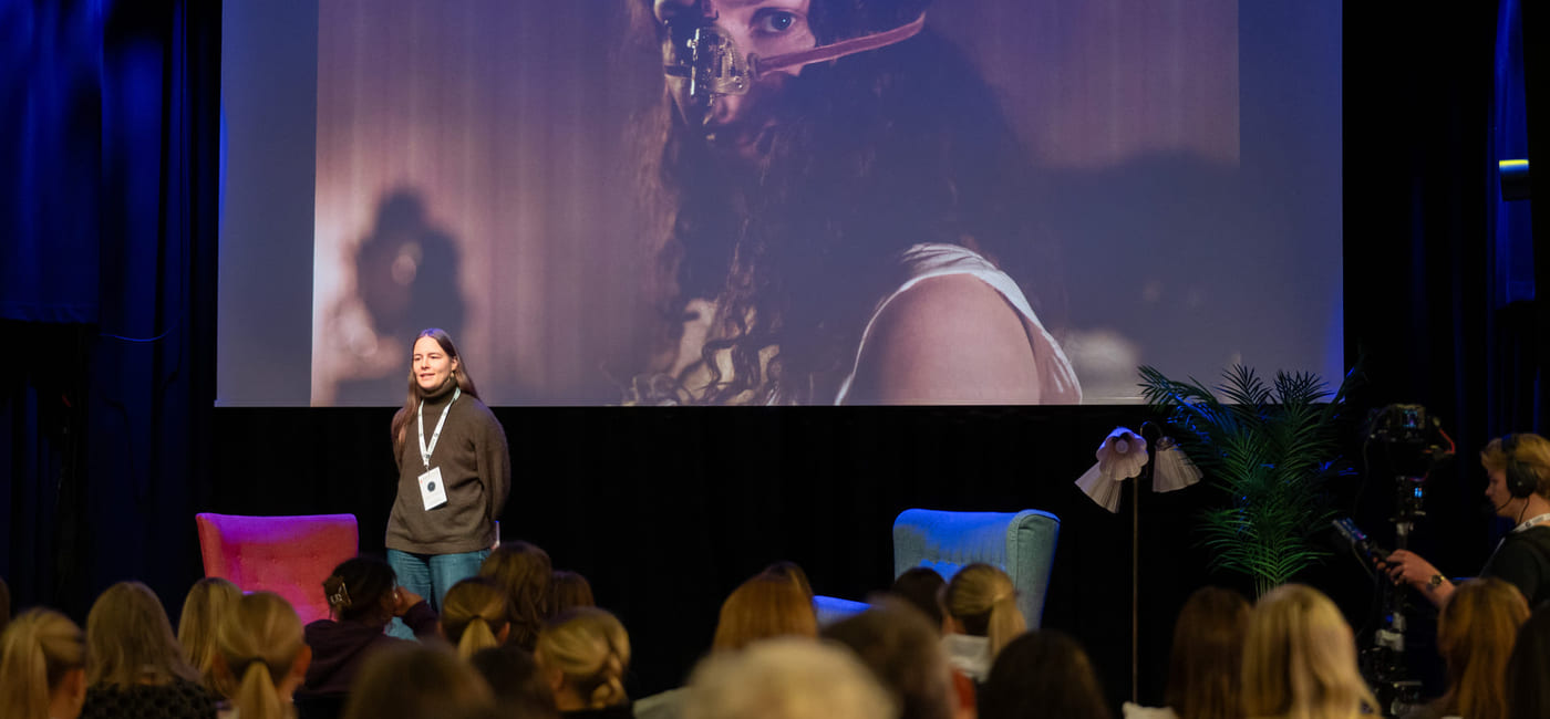 En kvinne står foran en storskjerm på en scene og snakker til et publikum.