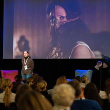 En kvinne står foran en storskjerm på en scene og snakker til et publikum.