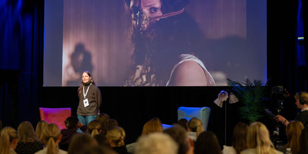 En kvinne står foran en storskjerm på en scene og snakker til et publikum.