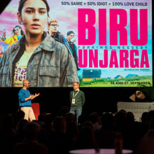 To menn står på en scene og snakker med hverandre foran en storskjerm.