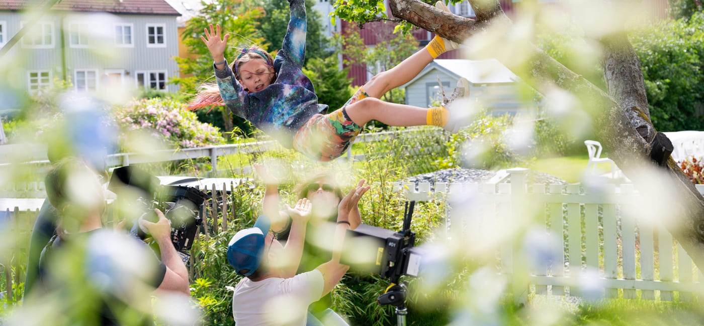 En jente faller fra et tre. To menn tar i mot henne og et kamera filmer.