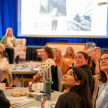 En gruppe publikummere sitter ved runde bord foran en scene. De ser bak seg og smiler.