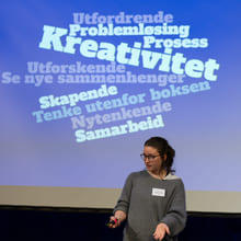 En kvinne med grå genser og briller står foran en skjerm hvor det er en ordsky. "Kreativitet" er størst.