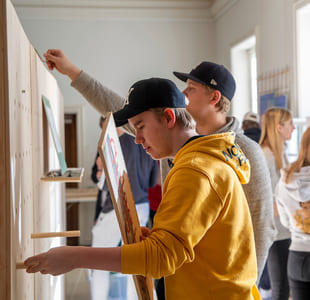 En gruppe elever henger opp noe på en vegg. en gutt i gul hettegenser og caps står fremst.