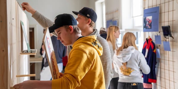 En gruppe elever henger opp noe på en vegg. en gutt i gul hettegenser og caps står fremst.