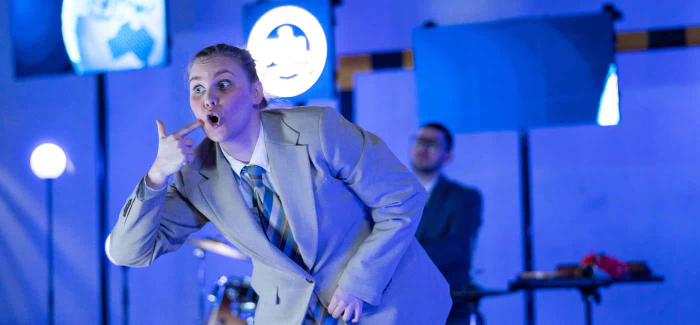 En skuespiller står med bøyd rygg på en scene, hun holder en finger til kinnet.