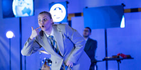 En skuespiller står med bøyd rygg på en scene, hun holder en finger til kinnet.