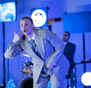 En skuespiller står med bøyd rygg på en scene, hun holder en finger til kinnet.