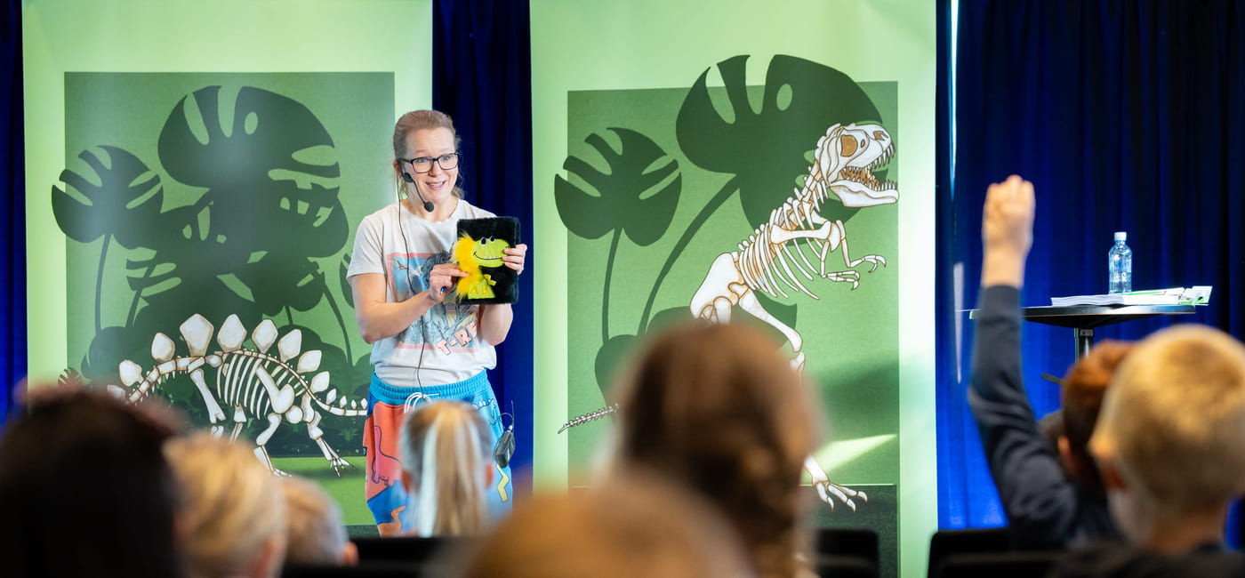 En kvinne står på en scene foran et publikum og holder en bok. Et barn rekker hånden i været.