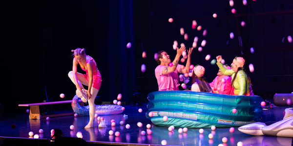 Fire skuespillere sitter i et barnebasseng og kaster baller i lufta, mens en femte tar på seg skoene.