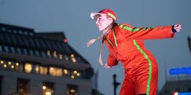 En person i rød kjeledress og blonde fletter lener seg utover. Det er mørkt, hun er opplyst av scenelys. Vi er ute.