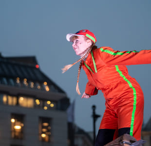 En person i rød kjeledress og blonde fletter lener seg utover. Det er mørkt, hun er opplyst av scenelys. Vi er ute.