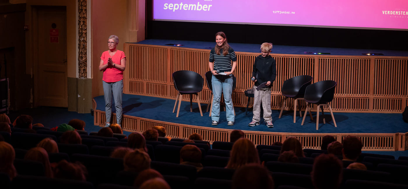 To barn og en kvinne står på en scene foran en storskjerm og prater til en kinosal me dpublikum.