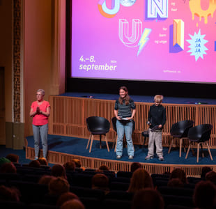 To barn og en kvinne står på en scene foran en storskjerm og prater til en kinosal me dpublikum.