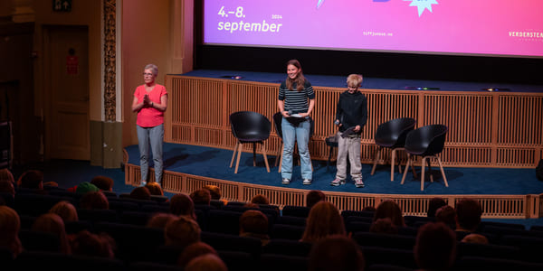 To barn og en kvinne står på en scene foran en storskjerm og prater til en kinosal me dpublikum.