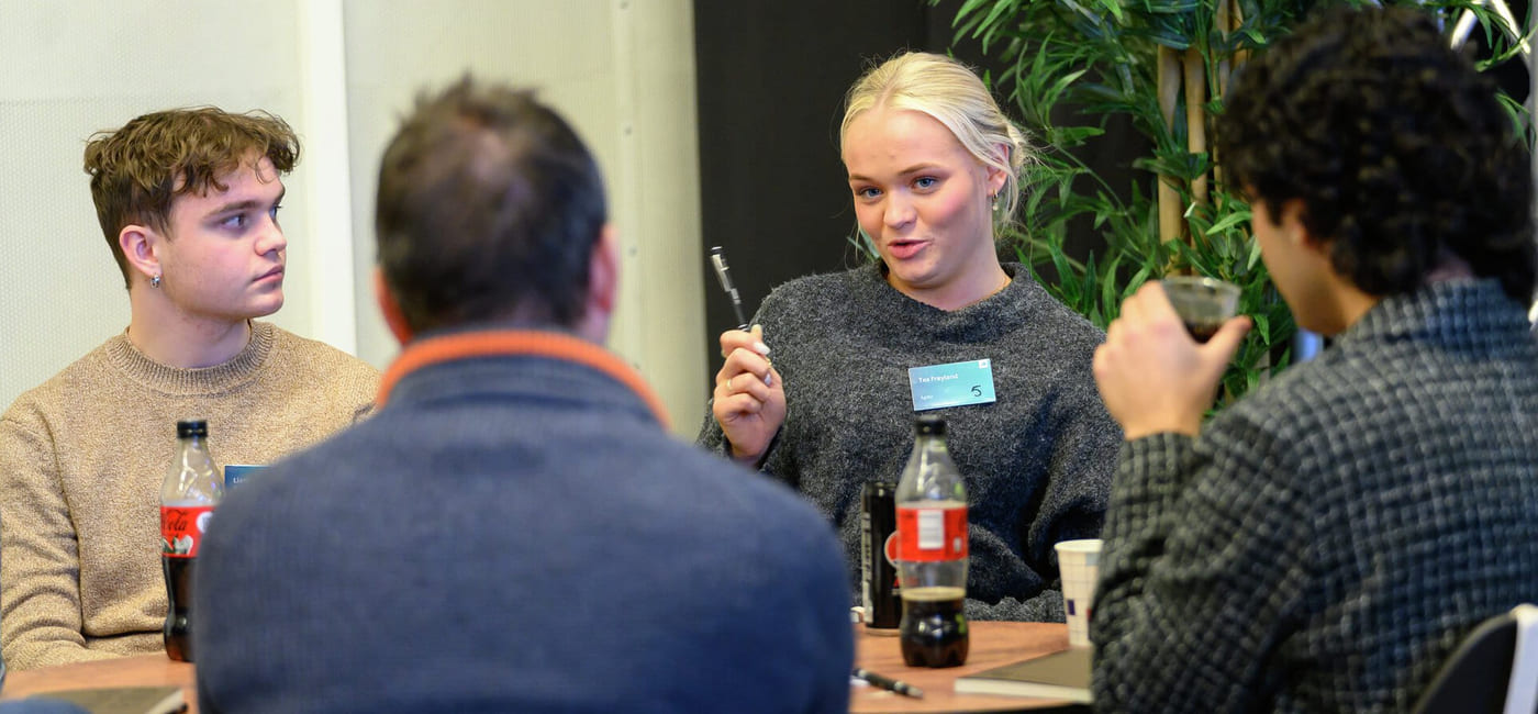 Fire personer sitter ved et bord. En ung kvinne snakker.