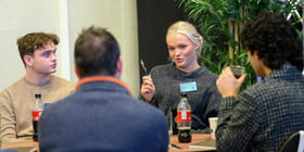 Fire personer sitter ved et bord. En ung kvinne snakker.