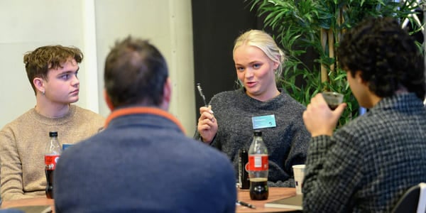 Fire personer sitter ved et bord. En ung kvinne snakker.