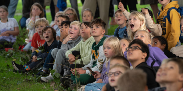 Flere barn sitter ved siden av hverandre på en gressplen, de måper og smiler mot noe utenfor bildet.