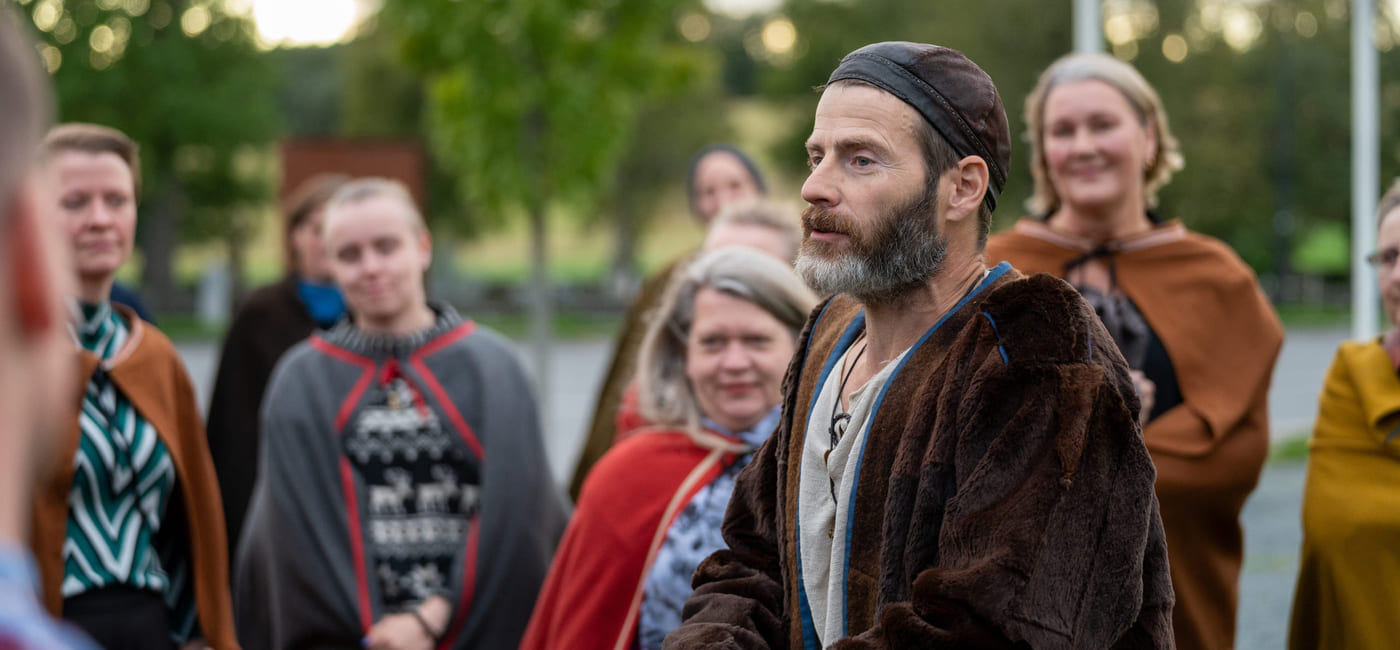 En mann i vikingdrakt snakker med en gruppe mennesker, som ser nysgjerrig på ham.