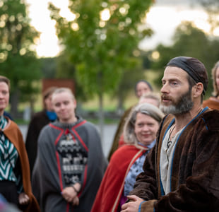 En mann i vikingdrakt snakker med en gruppe mennesker, som ser nysgjerrig på ham.