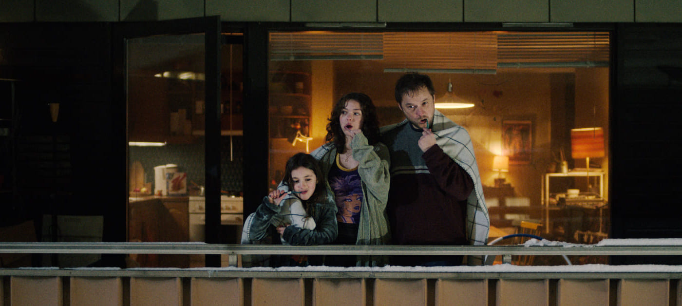 En mann og to unge jenter står på en balkong og pusser tennene sine. Alle har tepper over skuldrene sine.