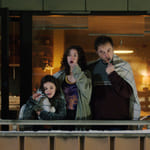 En mann og to unge jenter står på en balkong og pusser tennene sine. Alle har tepper over skuldrene sine.