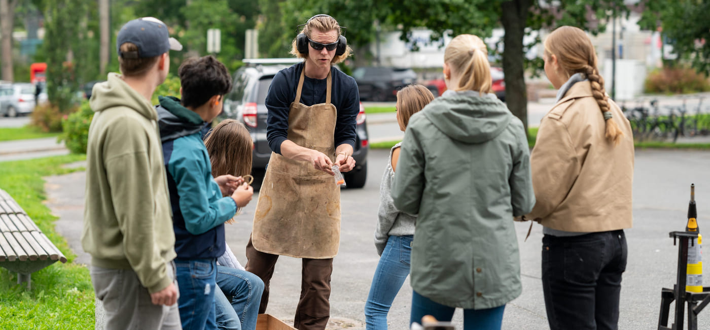 En gruppe ungdommer er samlet rundt en smed i en skolegård. Smeden har på seg verneutstyr,