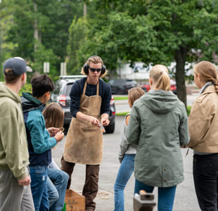 En gruppe ungdommer er samlet rundt en smed i en skolegård. Smeden har på seg verneutstyr,