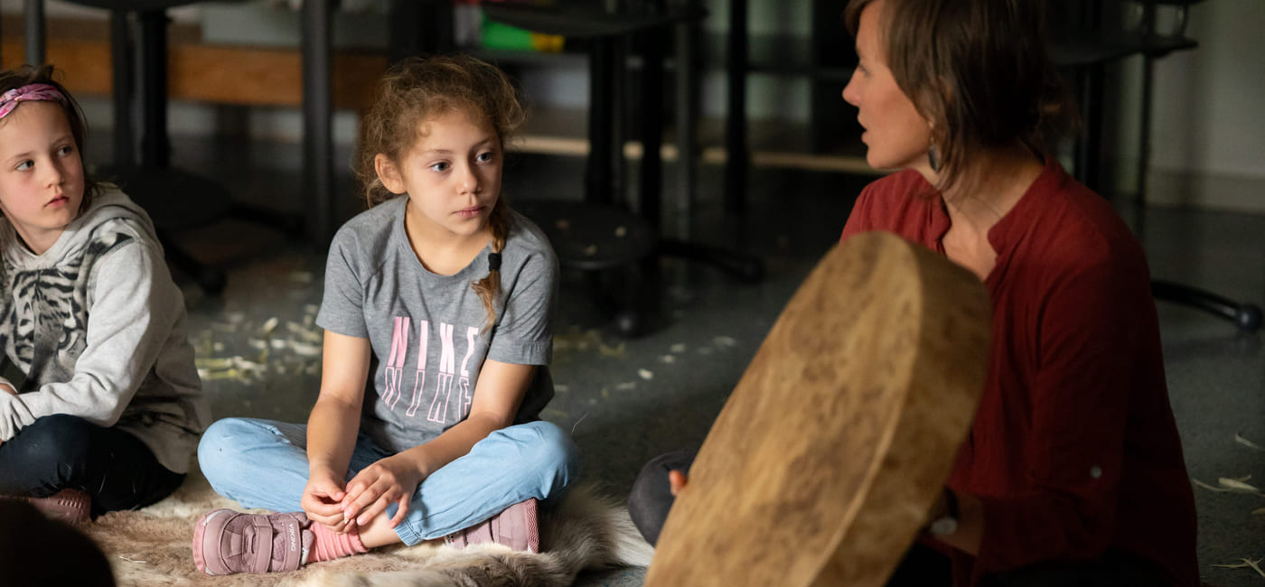 To jenter sitter på et gulv og ser på en tromme laget av skinn som en kvinne holder opp. Kvinnen sitter ved siden av jentene på gulvet.