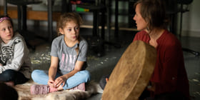 To jenter sitter på et gulv og ser på en tromme laget av skinn som en kvinne holder opp. Kvinnen sitter ved siden av jentene på gulvet.