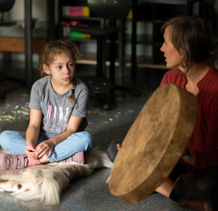 To jenter sitter på et gulv og ser på en tromme laget av skinn som en kvinne holder opp. Kvinnen sitter ved siden av jentene på gulvet.