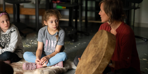 To jenter sitter på et gulv og ser på en tromme laget av skinn som en kvinne holder opp. Kvinnen sitter ved siden av jentene på gulvet.