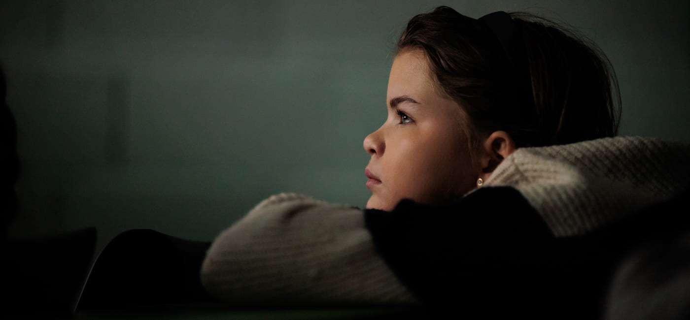 Et barn ser tenksomt mot noe utenfor bildet, med svakt lys i rommet.