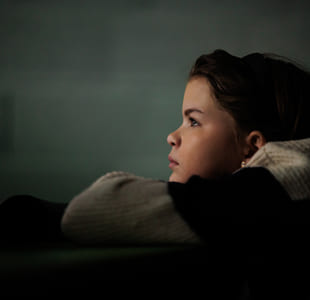 Et barn ser tenksomt mot noe utenfor bildet, med svakt lys i rommet.