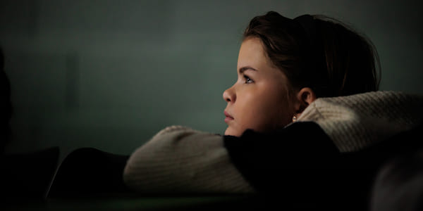 Et barn ser tenksomt mot noe utenfor bildet, med svakt lys i rommet.