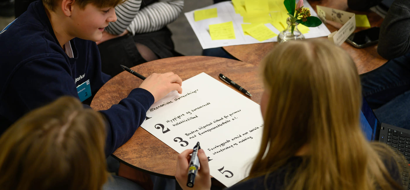 Studenter ved rundt bord jobber med en nummerert liste. Fargerike klisterlapper og en liten plante synlig.