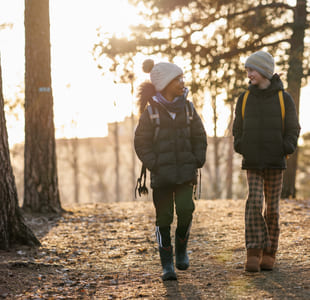 To jenter går ved siden av hverandre i et skoglandskap. De ser på hverandre og har på seg vinterklær. Det er lys fra en lav sol i bakgrunnen.