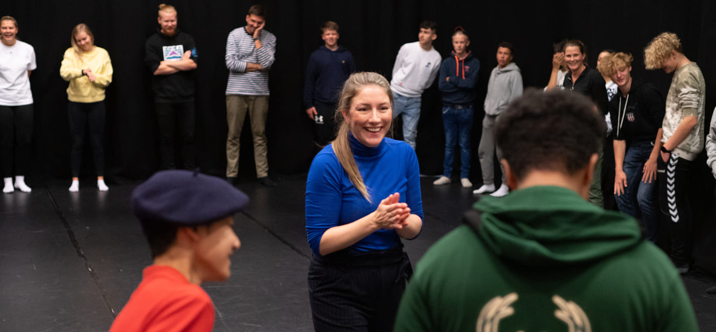 Kvinne i blå skjorte leder aktivitet for ulike grupper mennesker i teaterrommet, med tilskuere som ser på fra bakgrunnen.