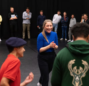 Kvinne i blå skjorte leder aktivitet for ulike grupper mennesker i teaterrommet, med tilskuere som ser på fra bakgrunnen.